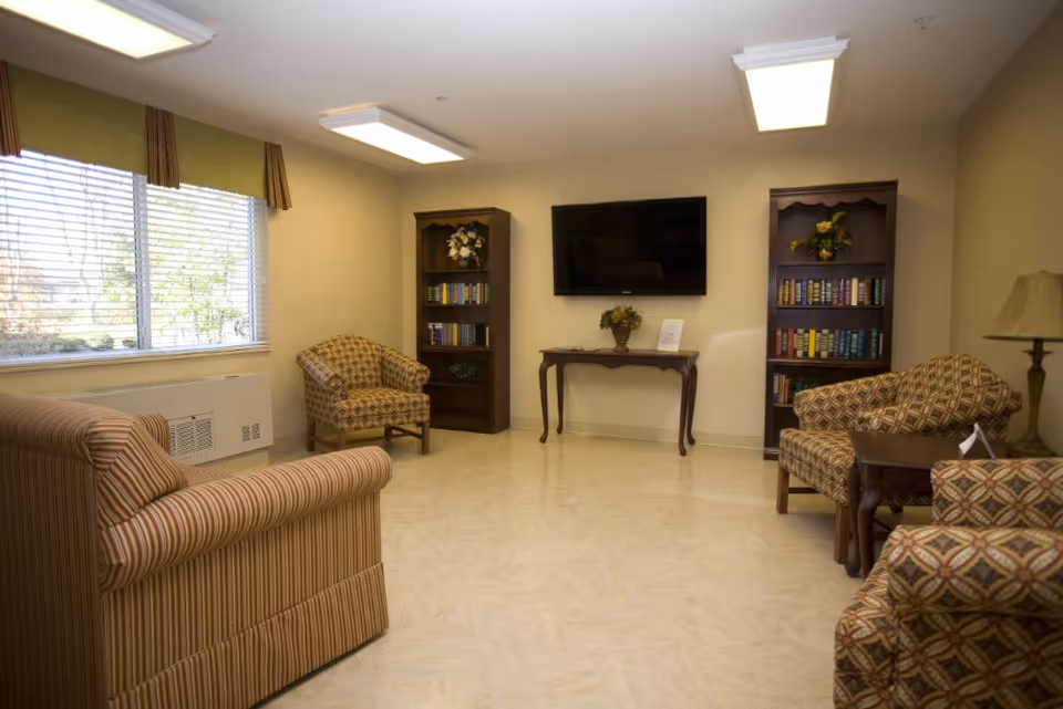 A furnished common room with upholstered chairs, bookshelves, a wall-mounted TV and a large window.