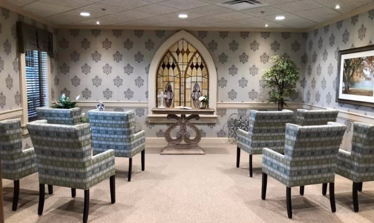 A small chapel or meditation room with patterned wallpaper, eight upholstered chairs arranged in two rows facing a decorative table with a stained glass window behind it. There is a potted plant in the corner and a framed landscape painting on the wall.