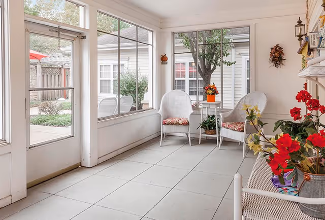 A bright indoor sunroom with large windows showing an outdoor garden area. The room features two white wicker chairs with floral cushions, a small white table with a potted plant, and a bench with colorful flowers in a vase. The walls are white, and there is a decorative wreath hanging on one wall.