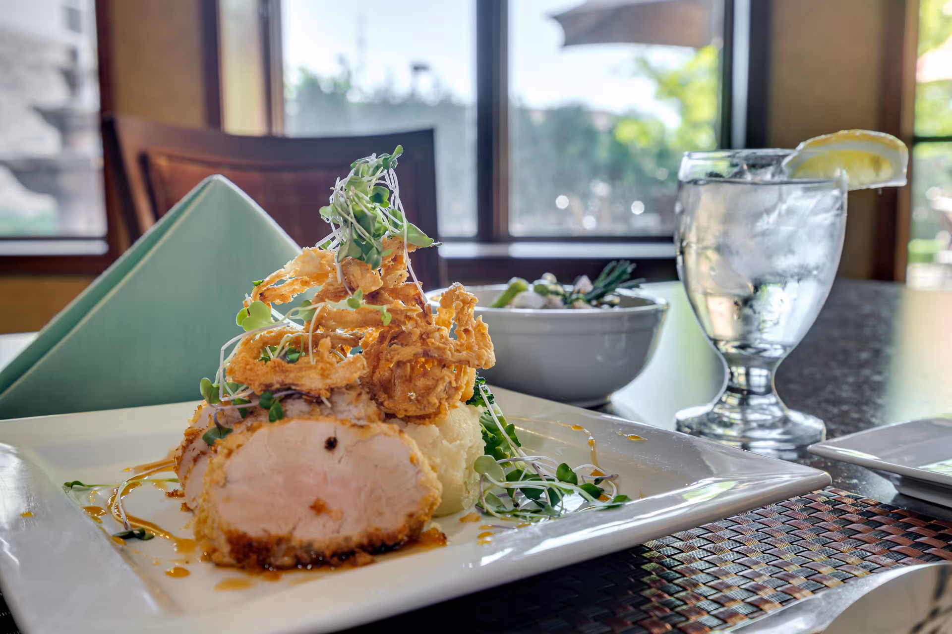 A plated meal featuring a breaded and cooked chicken breast topped with crispy fried onions and microgreens, served with mashed potatoes and a side of vegetables. A glass of water with a lemon wedge and a bowl of salad are also visible on the table near a window with natural light.