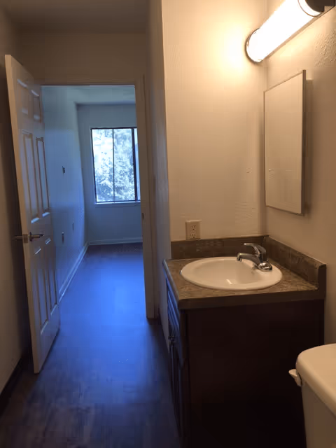 View of a small bathroom area with a sink, countertop, mirror, and light fixture on the right side. The bathroom opens into a hallway leading to a room with a window letting in natural light.