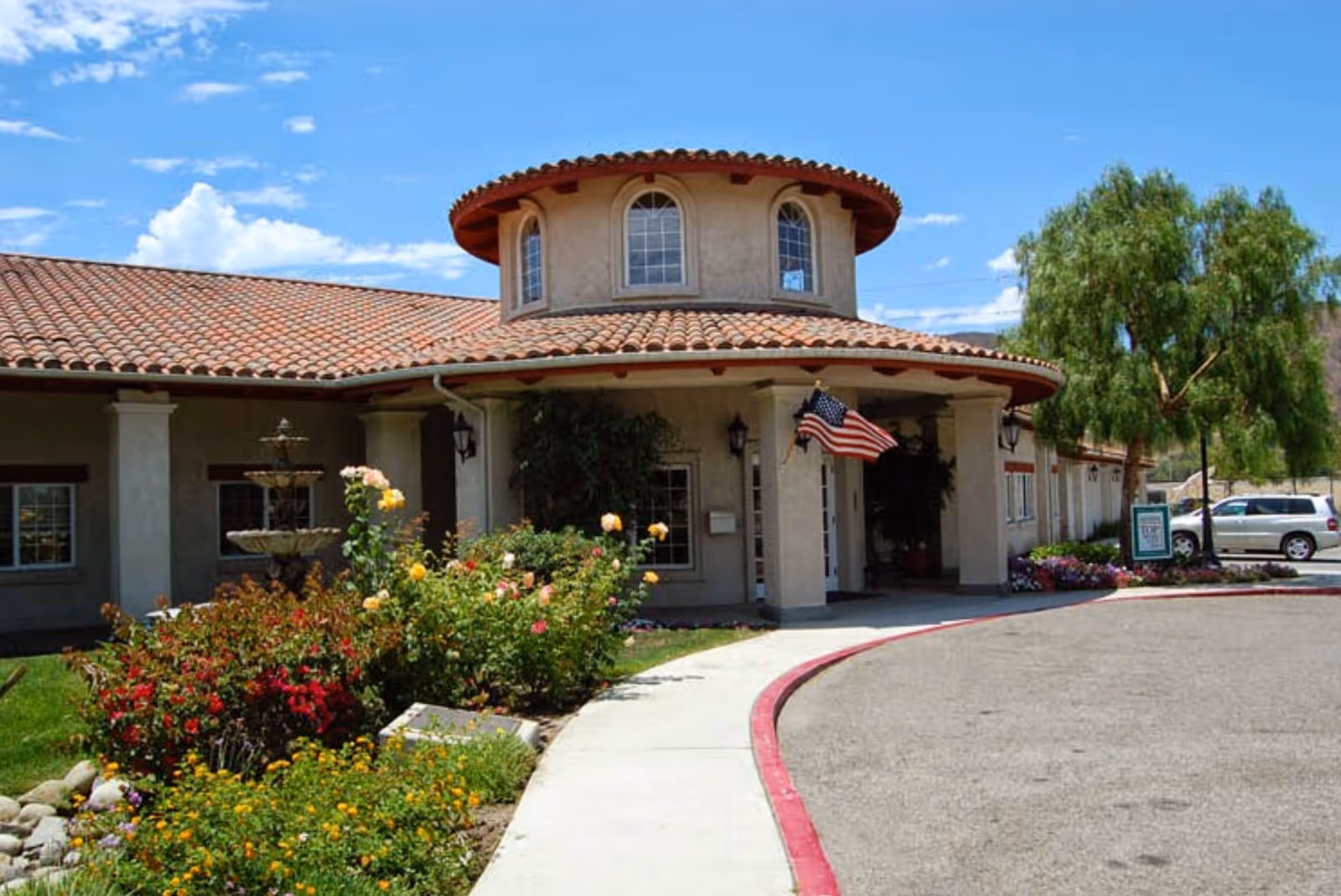 Exterior view of Silverado San Juan Capistrano Memory Care Community building with a tiled roof, a circular tower-like structure, an American flag near the entrance, a curved driveway, and landscaped garden with colorful flowers and a fountain.