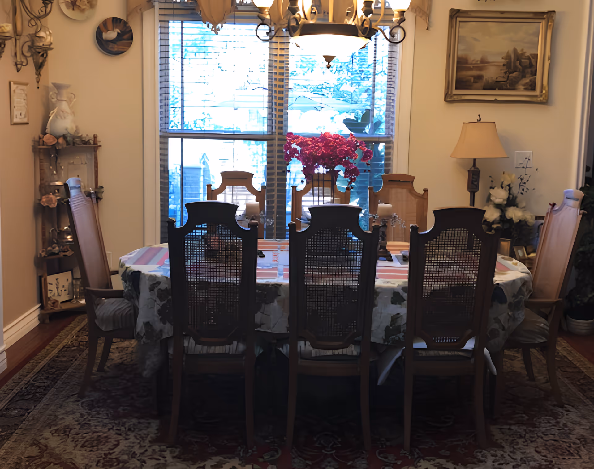 A dining room with a rectangular table covered with a floral tablecloth and surrounded by eight wooden chairs with woven backs. A vase with bright pink flowers is placed in the center of the table. The room has a large window with blinds letting in natural light, a chandelier hanging above the table, a floor lamp, and various decorative items on shelves and walls.