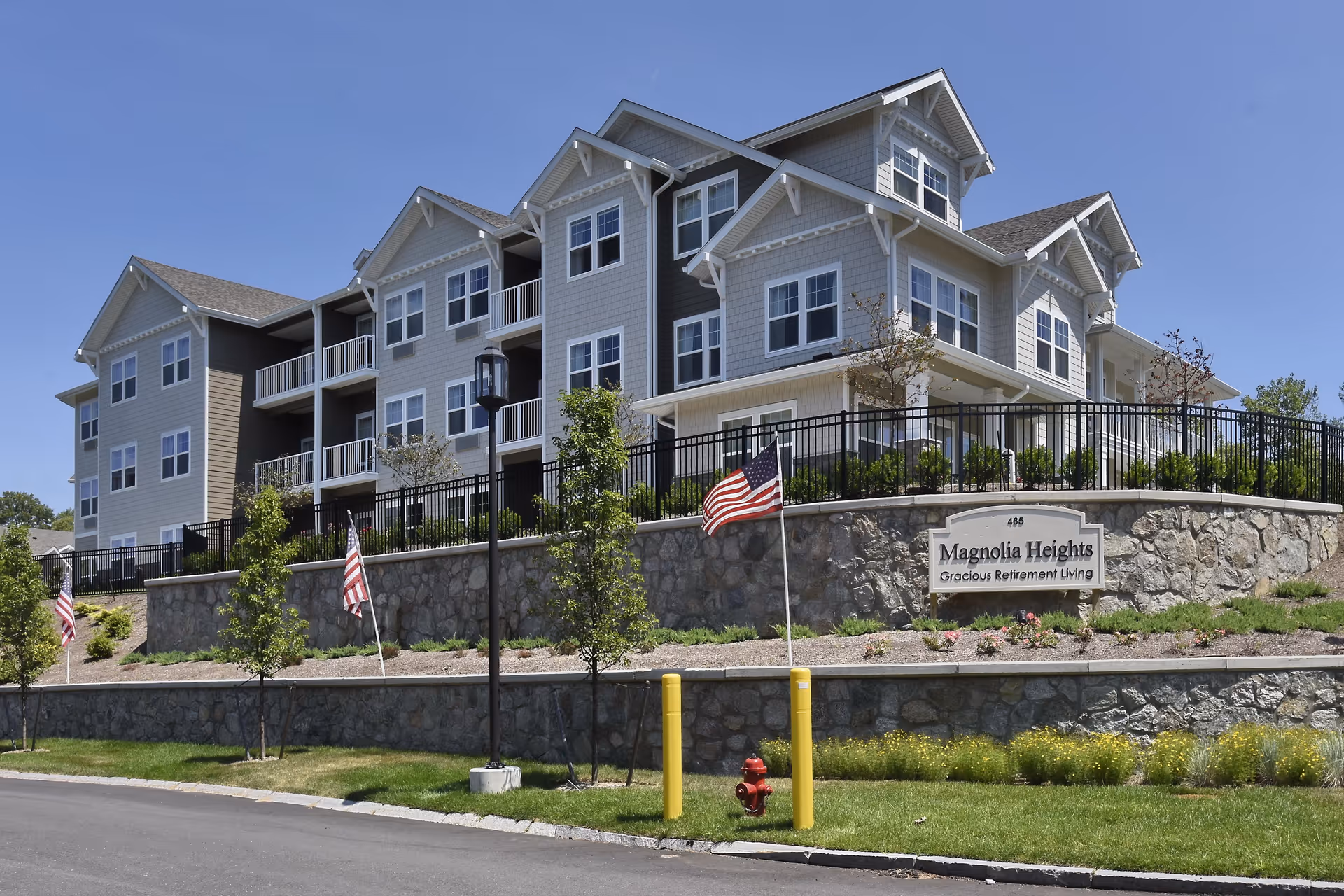 Exterior view of Magnolia Heights Gracious Retirement Living, a multi-story residential building with gray siding and white trim. The building is situated on a stone retaining wall with a landscaped area featuring small trees, shrubs, and American flags. A sign in front of the building displays the facility's name.