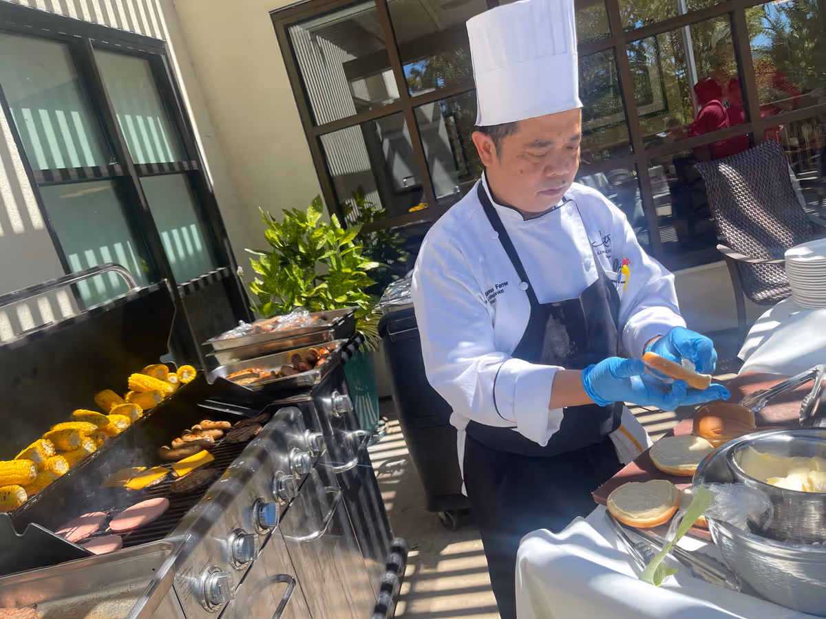 A chef in a white uniform and tall hat prepares burger buns beside a grill with corn, patties, and other foods at an outdoor buffet.