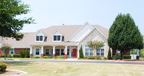 Front view of a single-story brick and stone senior living facility with a covered porch, American flag, red entry door, lawn and trees.