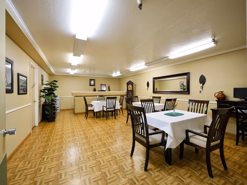 A dining room with several tables covered with white tablecloths and dark wooden chairs. The room has a parquet floor, beige walls, and fluorescent ceiling lights. There is a large mirror on one wall, framed pictures, a tall grandfather clock, a potted plant, and a small desk with a computer monitor and a decorative vase.