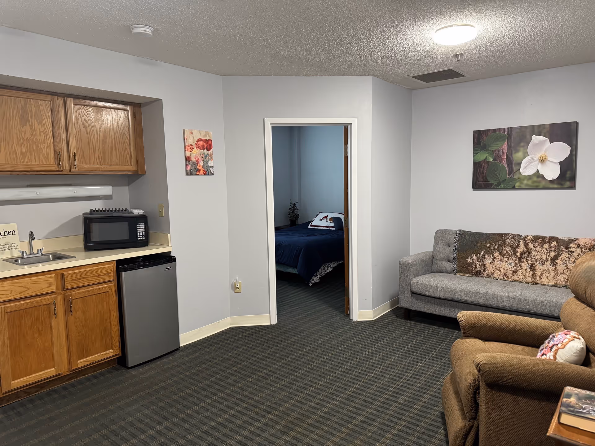 Interior view of a senior living facility suite showing a small kitchenette with wooden cabinets, a microwave, and a mini fridge on the left. In the center, there is a doorway leading to a bedroom with a bed covered in dark blue bedding. On the right side, there is a gray couch with a decorative pillow and a brown recliner chair with a small side table holding a book. The walls are light gray with floral artwork hanging on them, and the floor is carpeted with a dark patterned carpet.