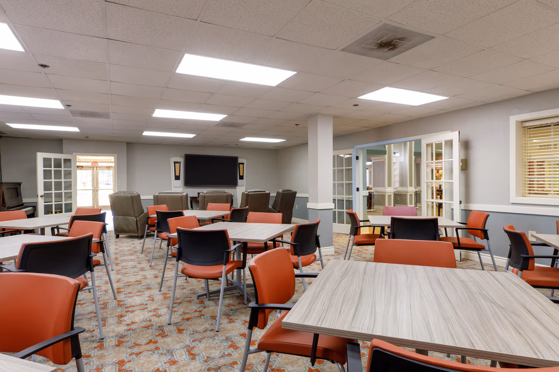 A spacious common room with multiple tables and chairs arranged for group seating. The chairs have orange and black seats with metal legs. The room has a patterned carpet floor, white ceiling tiles with fluorescent lighting, and light gray walls. At the far end, there are several recliner chairs facing a large flat-screen TV mounted on the wall. French doors lead to another area, and a window with blinds is visible on the right wall.