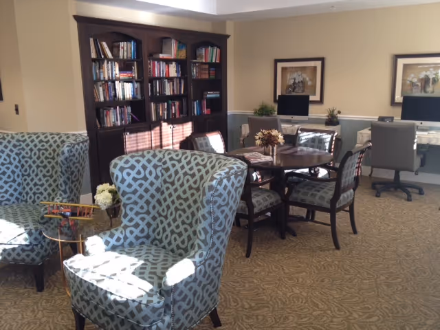 A cozy senior living common area with patterned armchairs, a round wooden table with chairs, a bookshelf filled with books, two computer desks with desktop computers, and framed artwork on the walls.