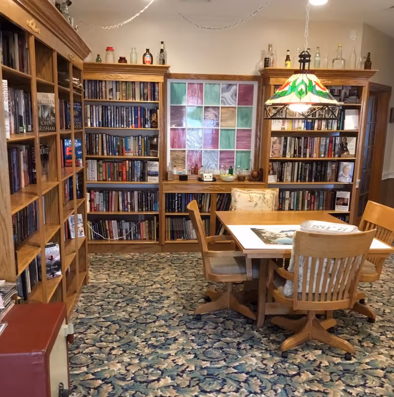 A cozy library room with wooden bookshelves filled with books along the walls. In the center, there is a wooden table with four wooden chairs on wheels. A colorful stained glass window is mounted on the wall above a lower bookshelf. A decorative hanging lamp with a green and red pattern illuminates the table. The floor is covered with a patterned carpet.
