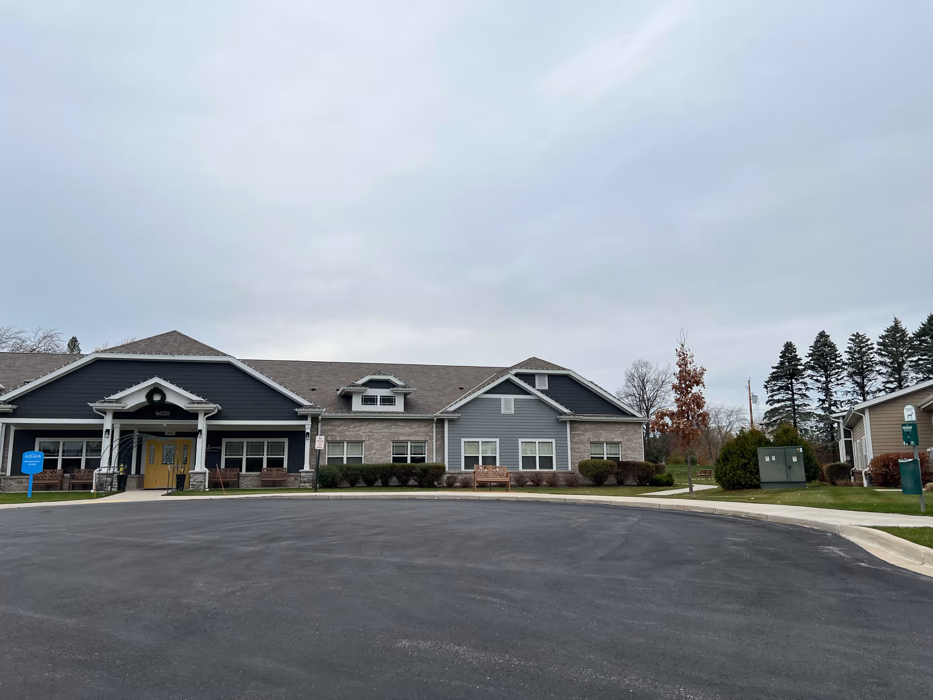 Exterior view of a single-story building with a dark blue and gray facade, a yellow entrance door, and a circular driveway in front. There are benches, bushes, and trees around the building under a cloudy sky.