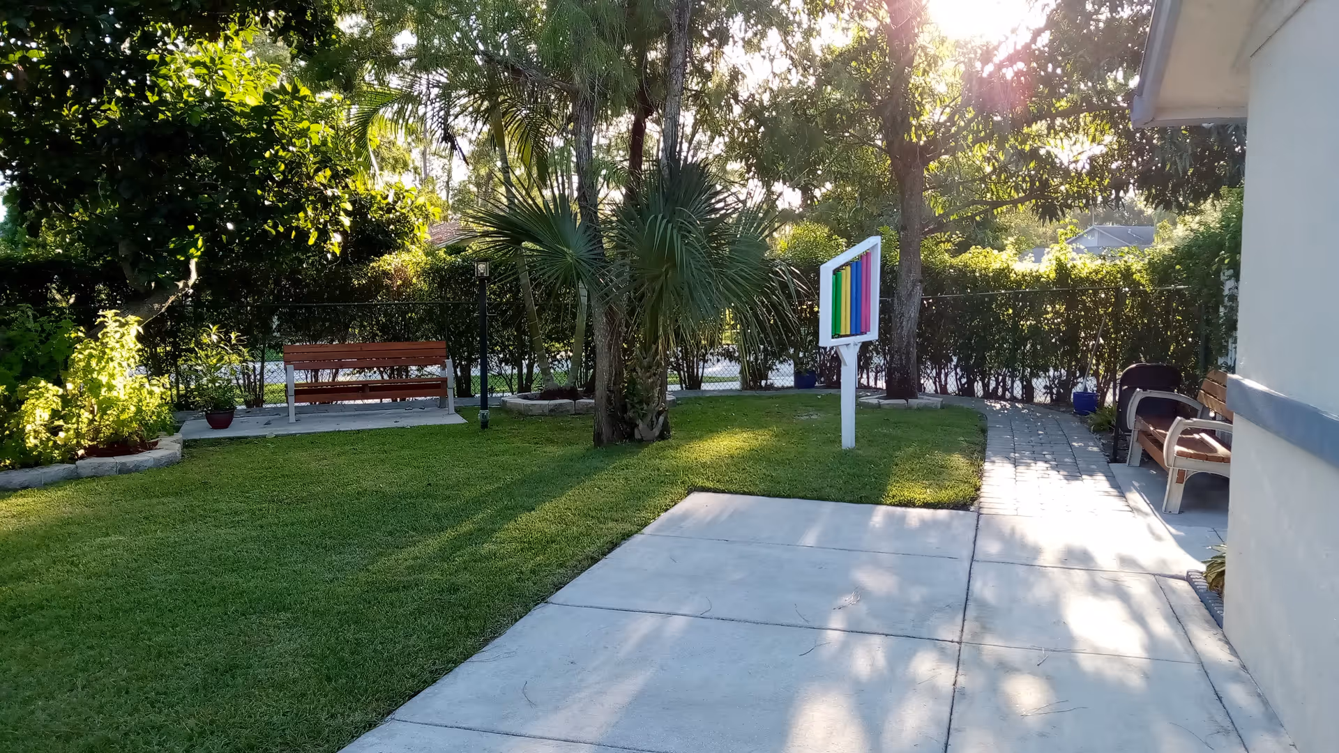 A sunny outdoor garden area with green grass, trees, and bushes. There are two wooden benches, one on a concrete slab and another near a building. A colorful vertical rainbow-striped sign is mounted on a white post in the grass. A paved walkway runs alongside the building on the right.