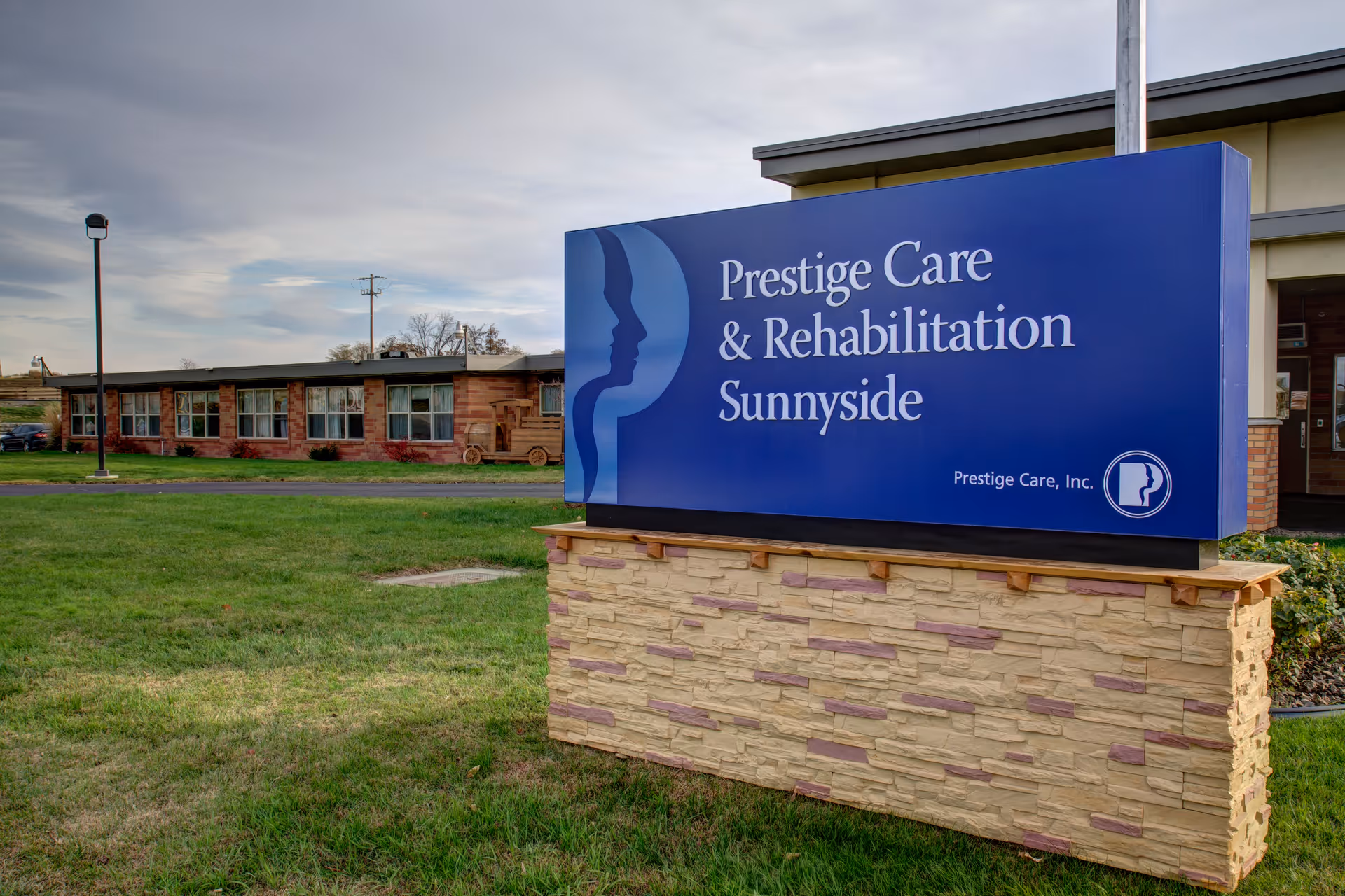 Outdoor view of Prestige Care & Rehabilitation Sunnyside facility sign on a stone base with the building and grassy area in the background under a cloudy sky.