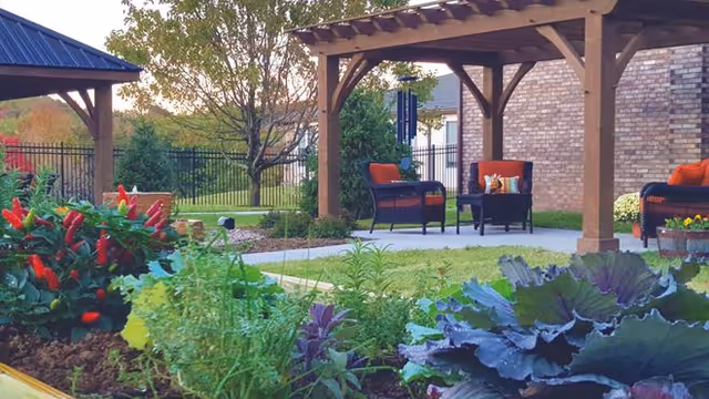 Outdoor garden area with a variety of plants and flowers in the foreground, and a wooden pergola with cushioned chairs and a small table underneath. A brick building and a black metal fence are visible in the background.