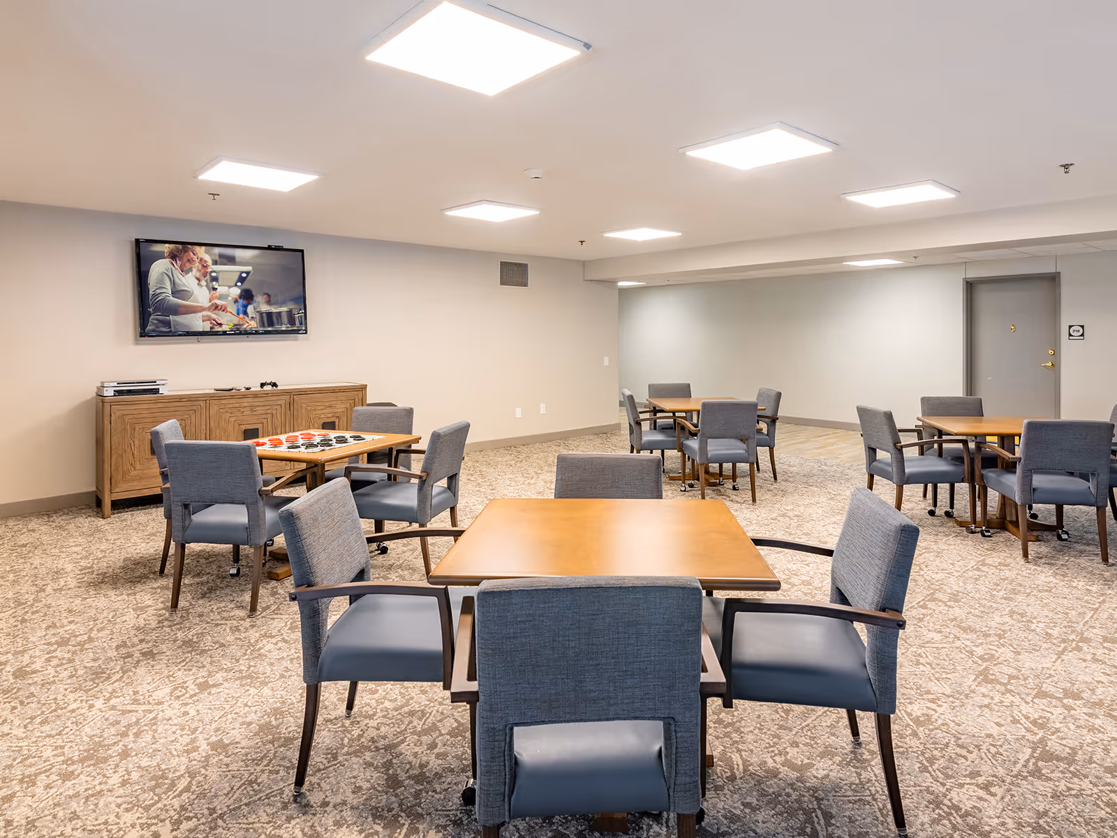 A spacious common room with several wooden tables and gray cushioned chairs arranged around them. A flat-screen TV is mounted on the wall above a wooden cabinet, displaying an image of two people cooking. The room has a carpeted floor and bright ceiling lights.