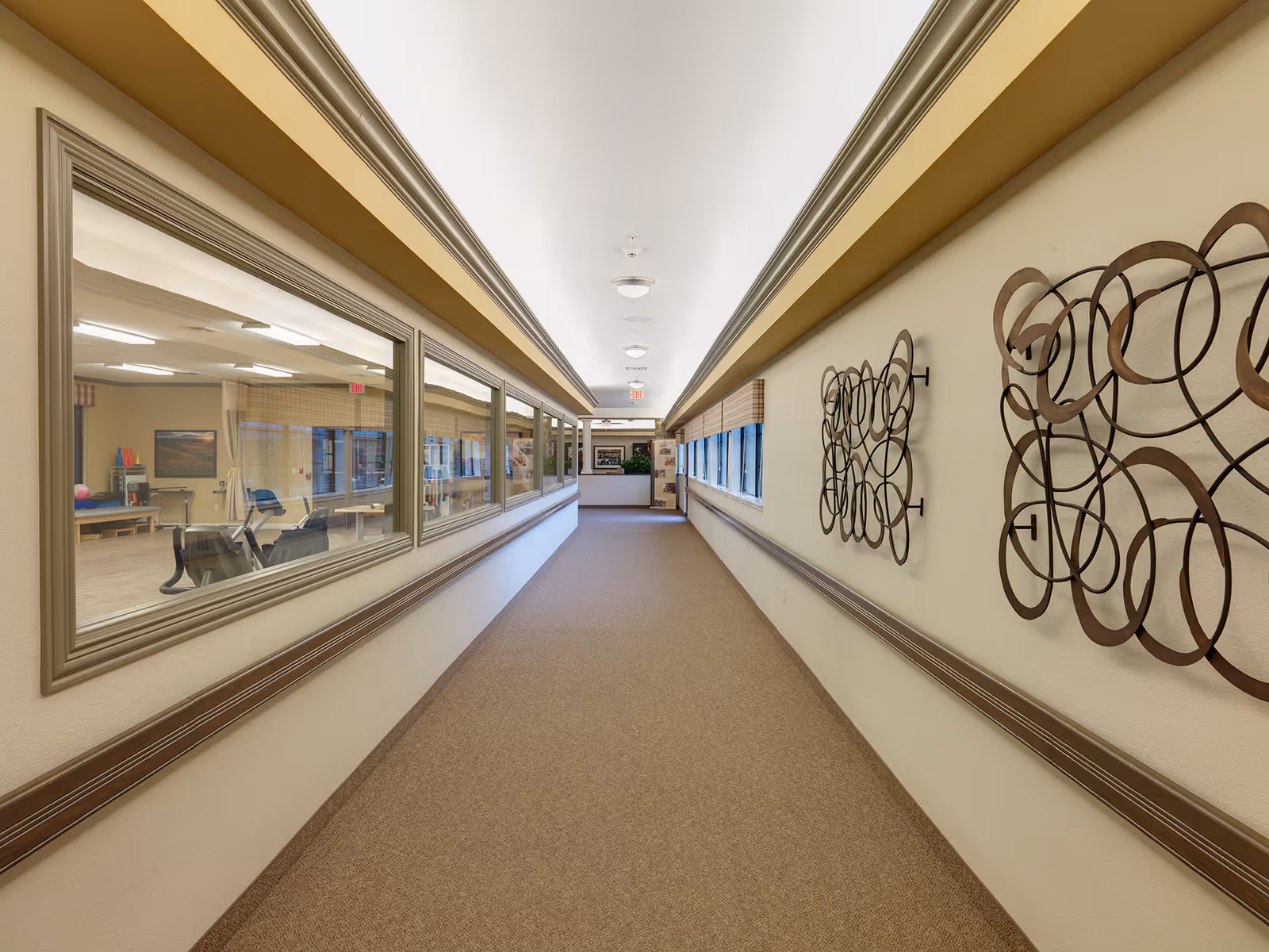 Long interior hallway with windows into activity rooms on the left and decorative metal wall art on the right.
