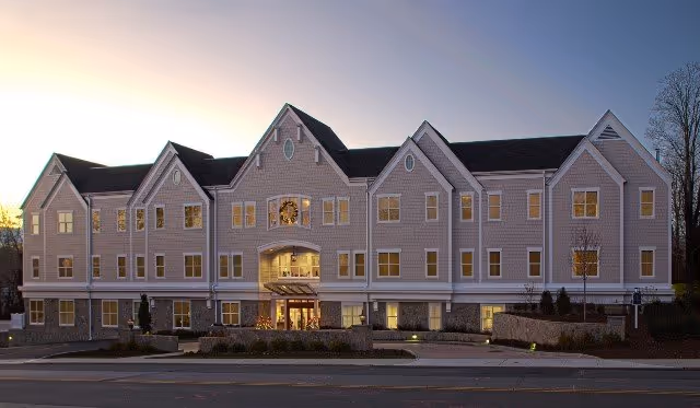 Exterior view of a large, multi-story senior living facility building at dusk with warm lights glowing from the windows and a decorated wreath above the main entrance.