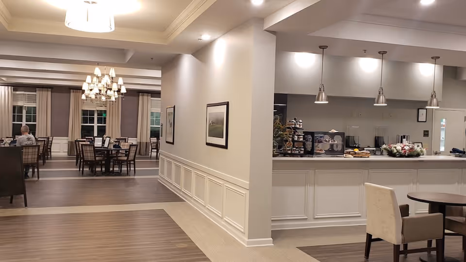Interior view of a senior living community dining area with multiple tables and chairs, a person sitting at one table, chandeliers hanging from the ceiling, and a counter area with pendant lights and various items on display.
