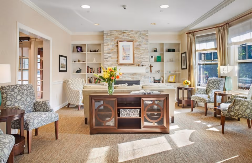 A bright and cozy living room with patterned armchairs, a beige sofa, a wooden console table with a vase of flowers, built-in white shelves, and a stone fireplace with a framed picture above it. Large windows with curtains allow natural light to fill the room.