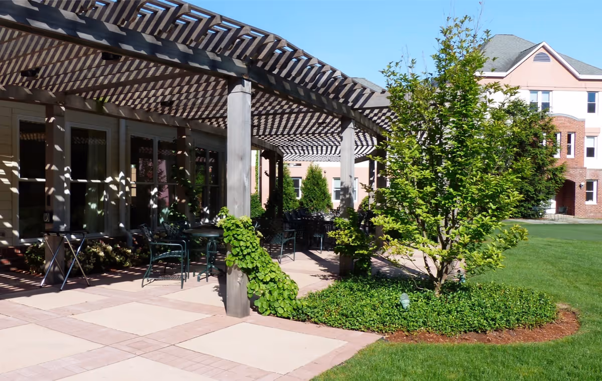 Outdoor patio area with a wooden pergola casting shadows over tables and chairs. There is a small tree surrounded by greenery and a well-maintained lawn. In the background, a multi-story building with brick and light-colored siding is visible under a clear blue sky.