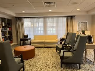 A cozy living room area with a yellow sofa against a window with curtains, four gray armchairs arranged around a round orange ottoman, a bookshelf on the left, and a small table with a lamp on the right.