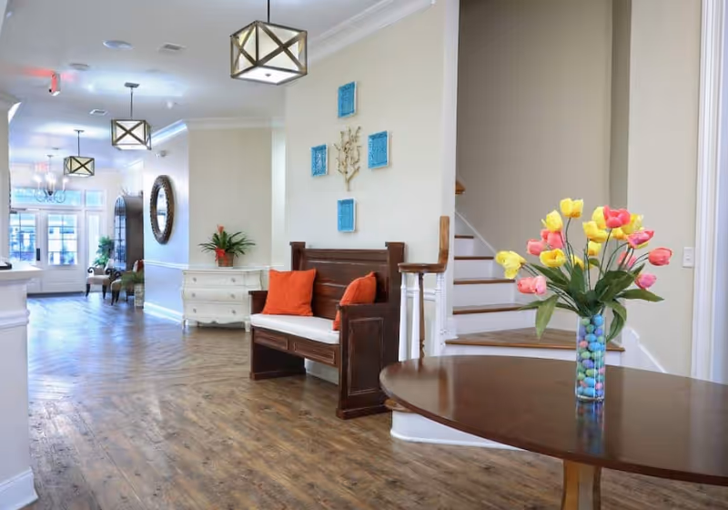 Bright, welcoming senior living lobby with wood floors, a bench with orange pillows, a round table holding a vase of tulips, and a staircase.