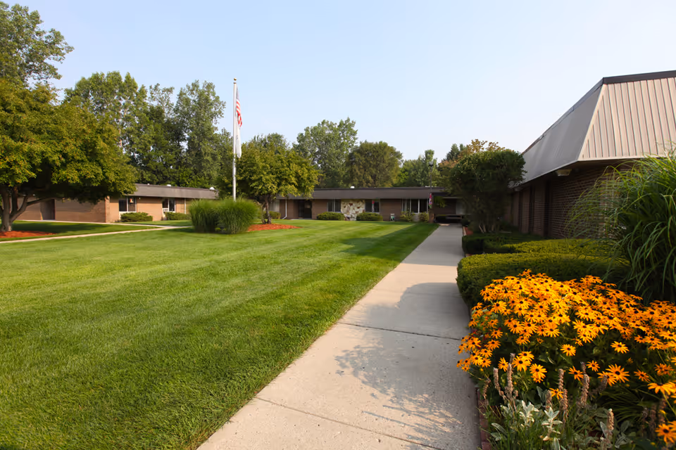 A well-maintained outdoor area of a senior living facility with a concrete walkway, green lawn, trees, and a flower bed with bright yellow flowers. The building is single-story with a brick exterior and a flagpole with an American flag in the center of the lawn.