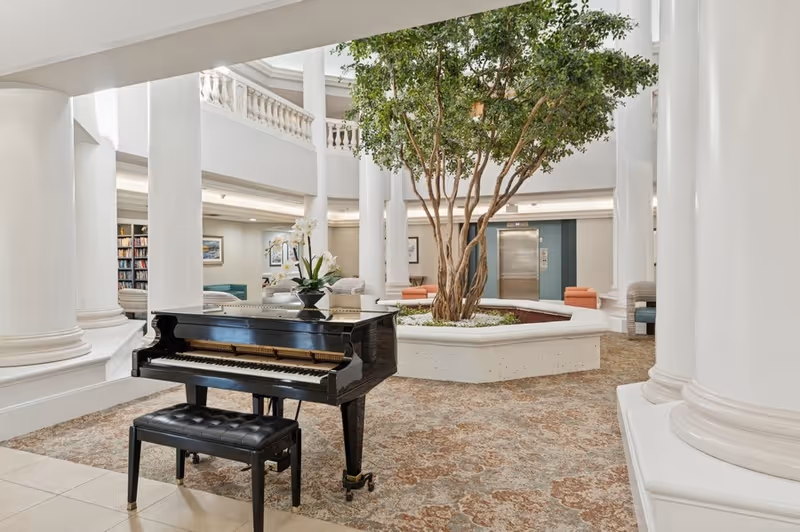 A spacious and elegant interior common area featuring a black grand piano with a matching bench, surrounded by large white columns. In the center, there is a large planter with a tall leafy tree. Comfortable seating areas and bookshelves are visible in the background, along with an elevator door.