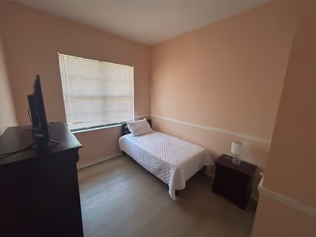 A small bedroom with a single bed covered in a white quilt and a white pillow. There is a window with closed blinds on the left wall, a dark wooden dresser with a TV on top near the window, and a dark wooden nightstand with a white lamp on the right side of the bed. The walls are painted light peach with a white chair rail molding.