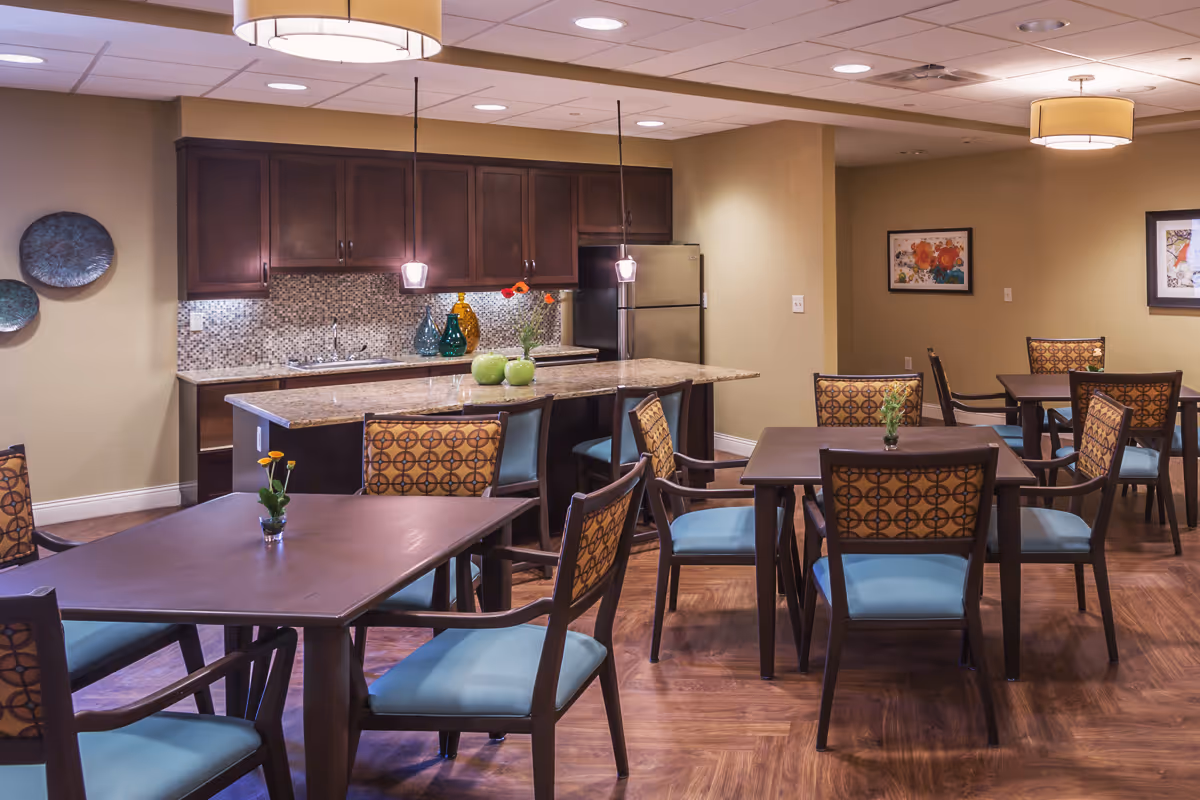 Dining room with several tables and patterned chairs arranged around a central kitchen island with pendant lights and a refrigerator in the background.
