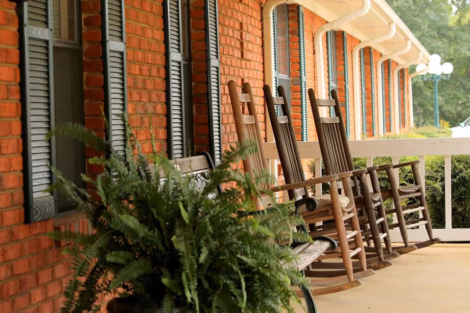 A porch at Magnolia New Albany featuring several wooden rocking chairs lined up against a brick wall, with green ferns in the foreground.