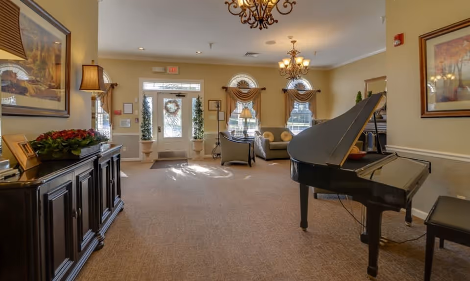 A spacious and well-lit living room area with beige walls and carpeted floor. The room features a black grand piano with a matching bench on the right side, two chandeliers hanging from the ceiling, a dark wooden cabinet with a lamp and flower arrangement on the left, and a seating area with a sofa and armchair near the windows. The entrance door is visible at the far end with decorative plants and a wreath on the door.