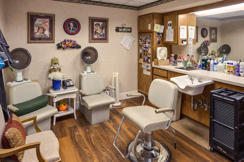 Small hair-salon room with styling chairs, hooded dryers, a sink and a countertop filled with hair products and cabinets.