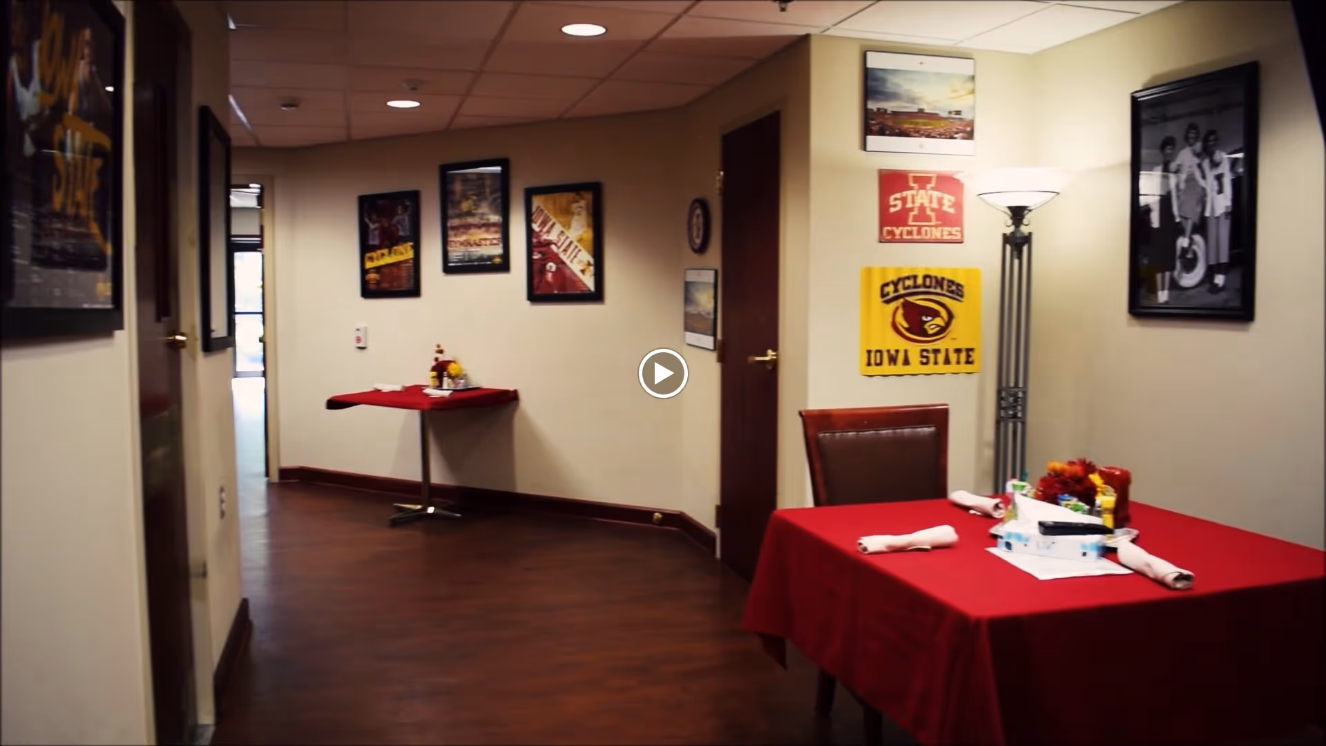 Interior hallway and small dining area with a table covered in a red tablecloth, set with napkins, condiments, and a flower centerpiece. The walls are decorated with framed pictures and posters, including Iowa State Cyclones memorabilia. A floor lamp is positioned near the table.