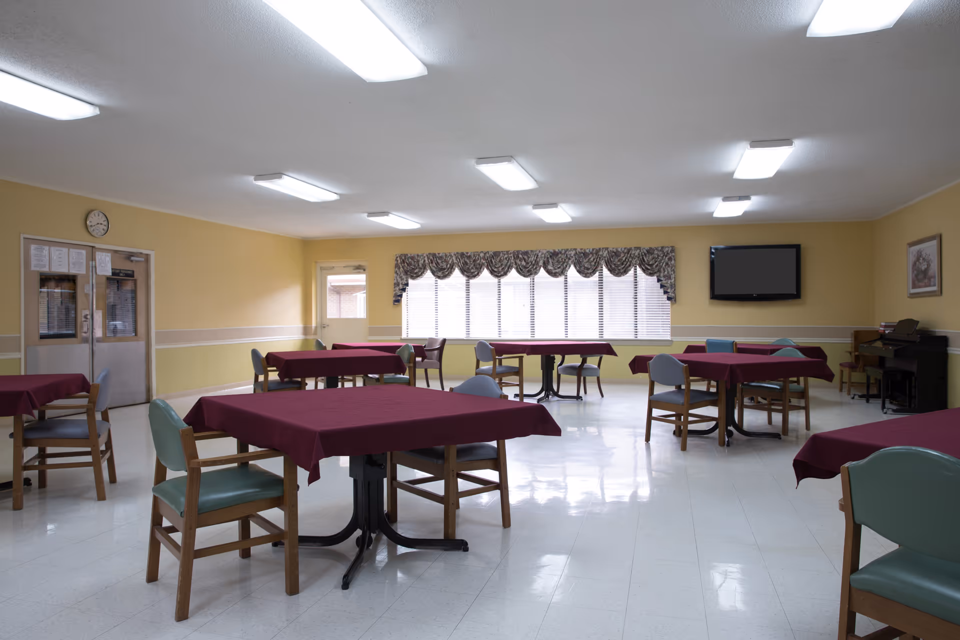 Spacious dining/activity room with multiple tables covered in maroon tablecloths and chairs, a wall-mounted TV, and a large window with valance.