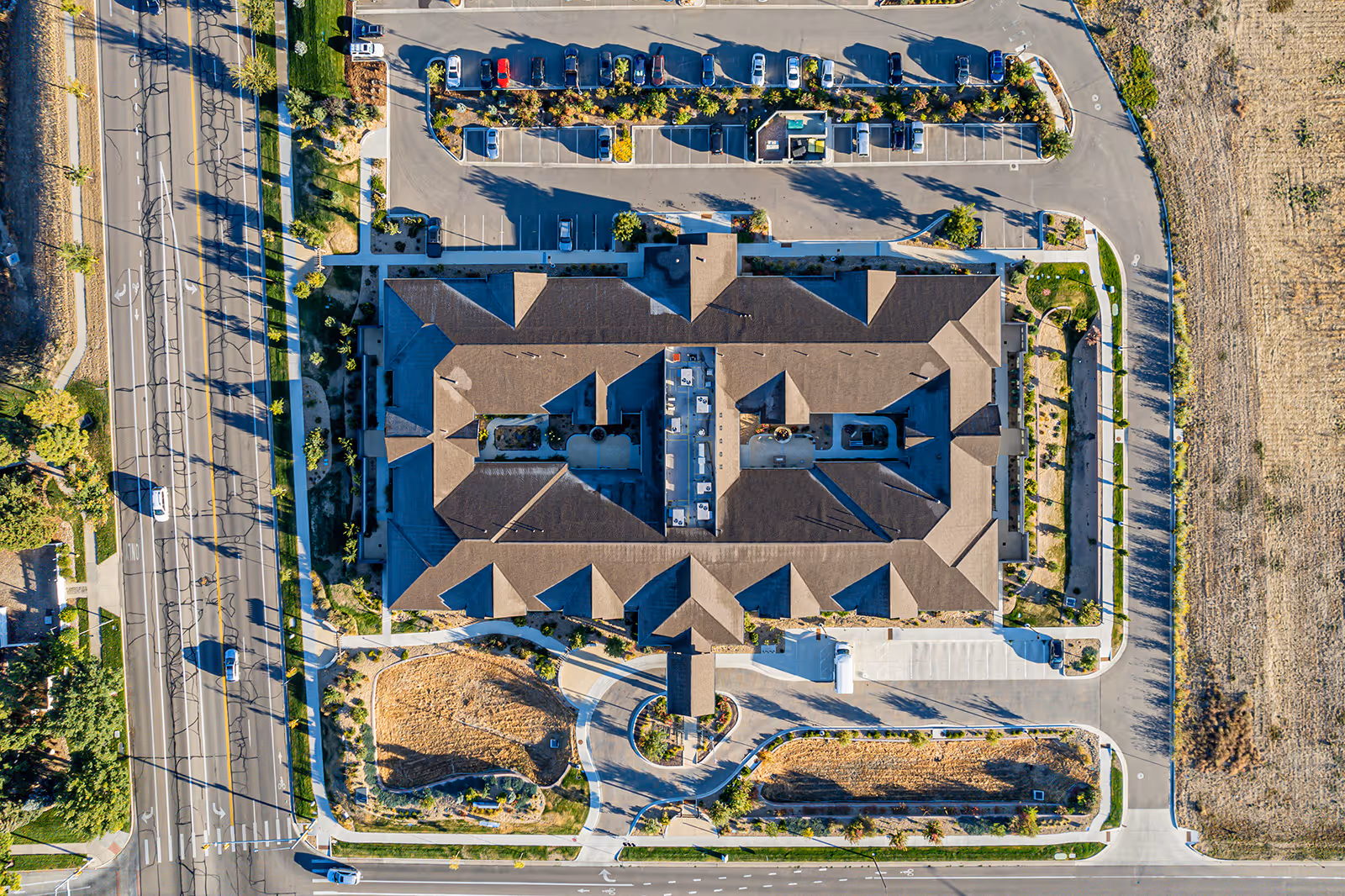 Aerial view of a large building with a brown roof surrounded by parking lots, roads, and landscaped areas. The building is situated in a developed area with some open land nearby.