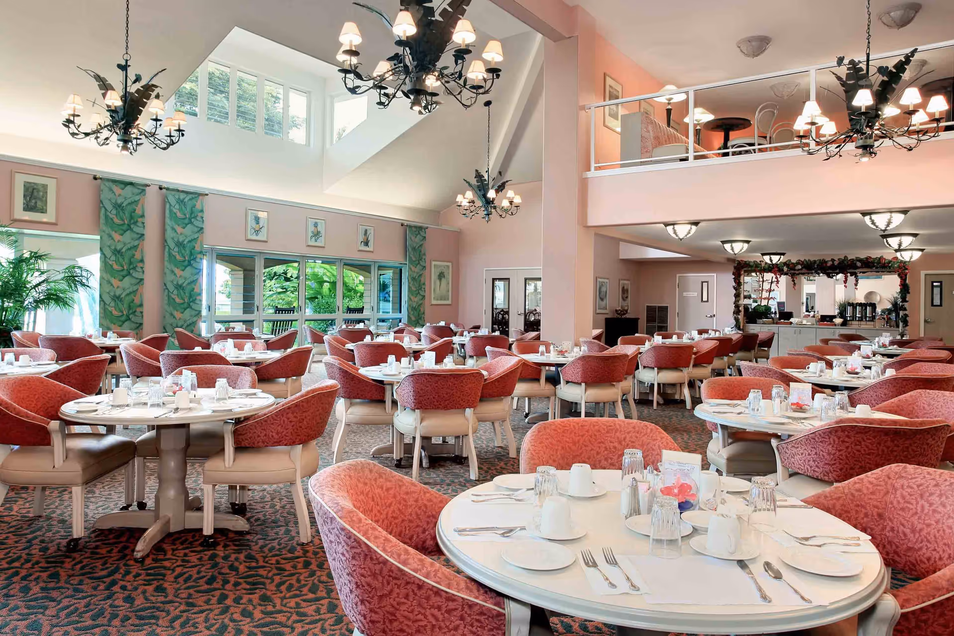 A spacious dining room with multiple round tables set with white plates, cups, utensils, and glasses. The room features pink walls, large windows with green leafy curtains, and several ornate chandeliers hanging from a high ceiling. There is a mezzanine level with seating visible above, and the room is well-lit with natural light and ceiling lights.