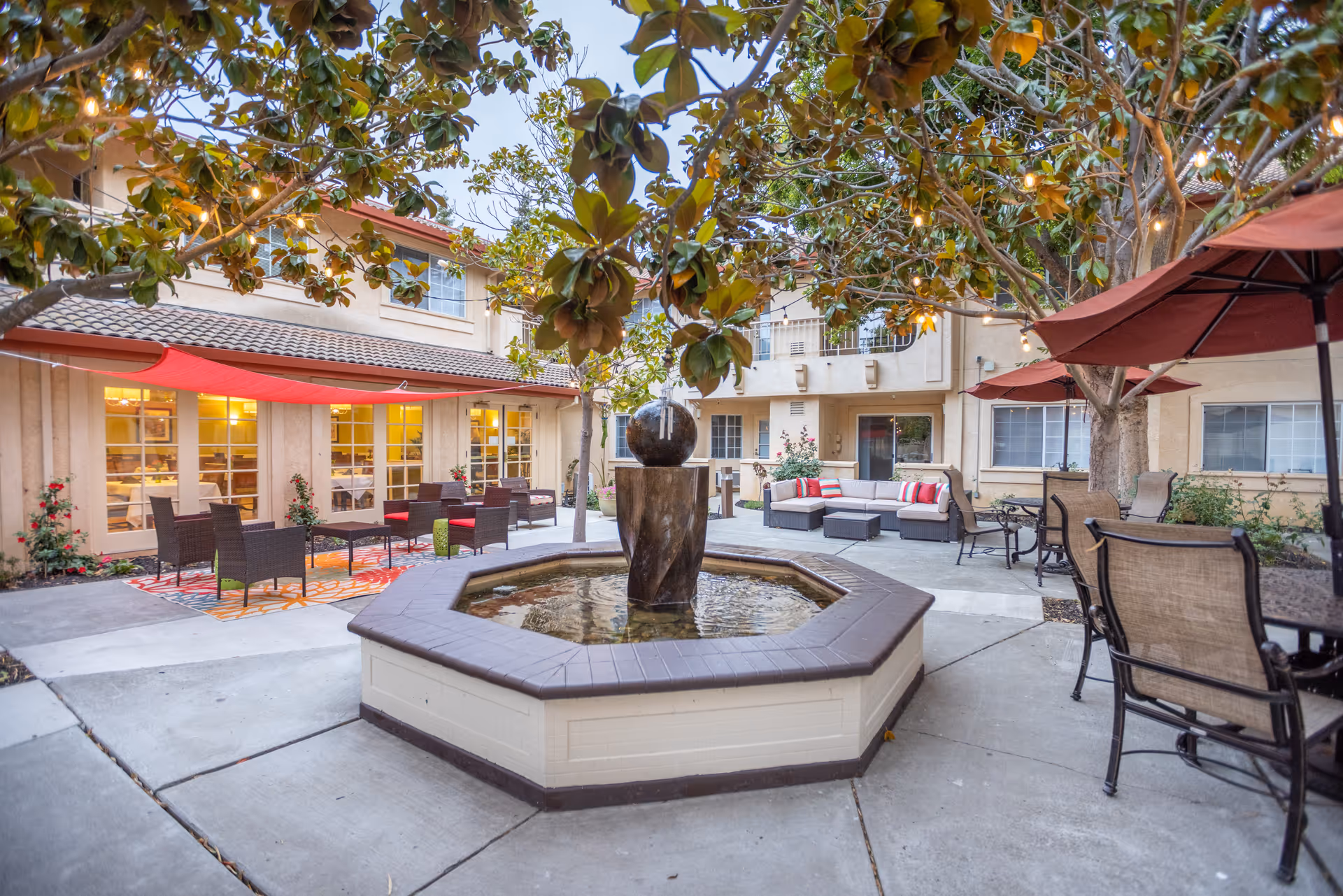 Outdoor courtyard area at Cogir of North Bay Senior Living featuring a central octagonal water fountain, surrounded by patio seating including cushioned chairs, tables with umbrellas, and a sofa set. Trees with hanging string lights provide shade and ambiance, with the building's windows and doors visible in the background.