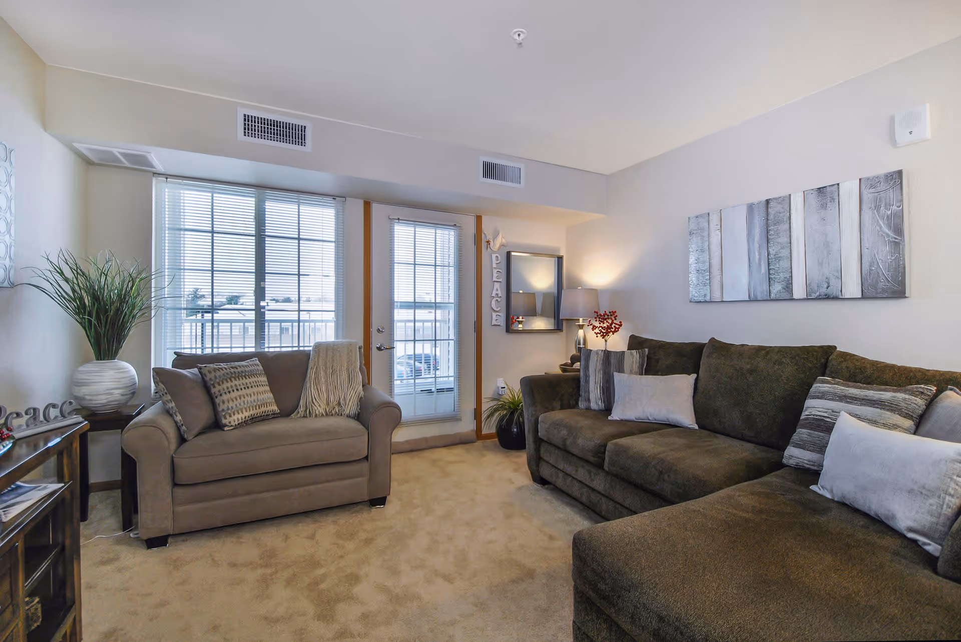 A cozy living room with a large dark green sectional sofa adorned with multiple pillows on the right and a smaller beige loveseat with pillows and a throw blanket on the left. There is a window and a glass door with blinds letting in natural light behind the loveseat. The walls are light-colored with a modern abstract painting above the sectional and a mirror with a lamp and decorative items on a small table in the corner. A potted plant sits on a wooden side table on the left side of the room.