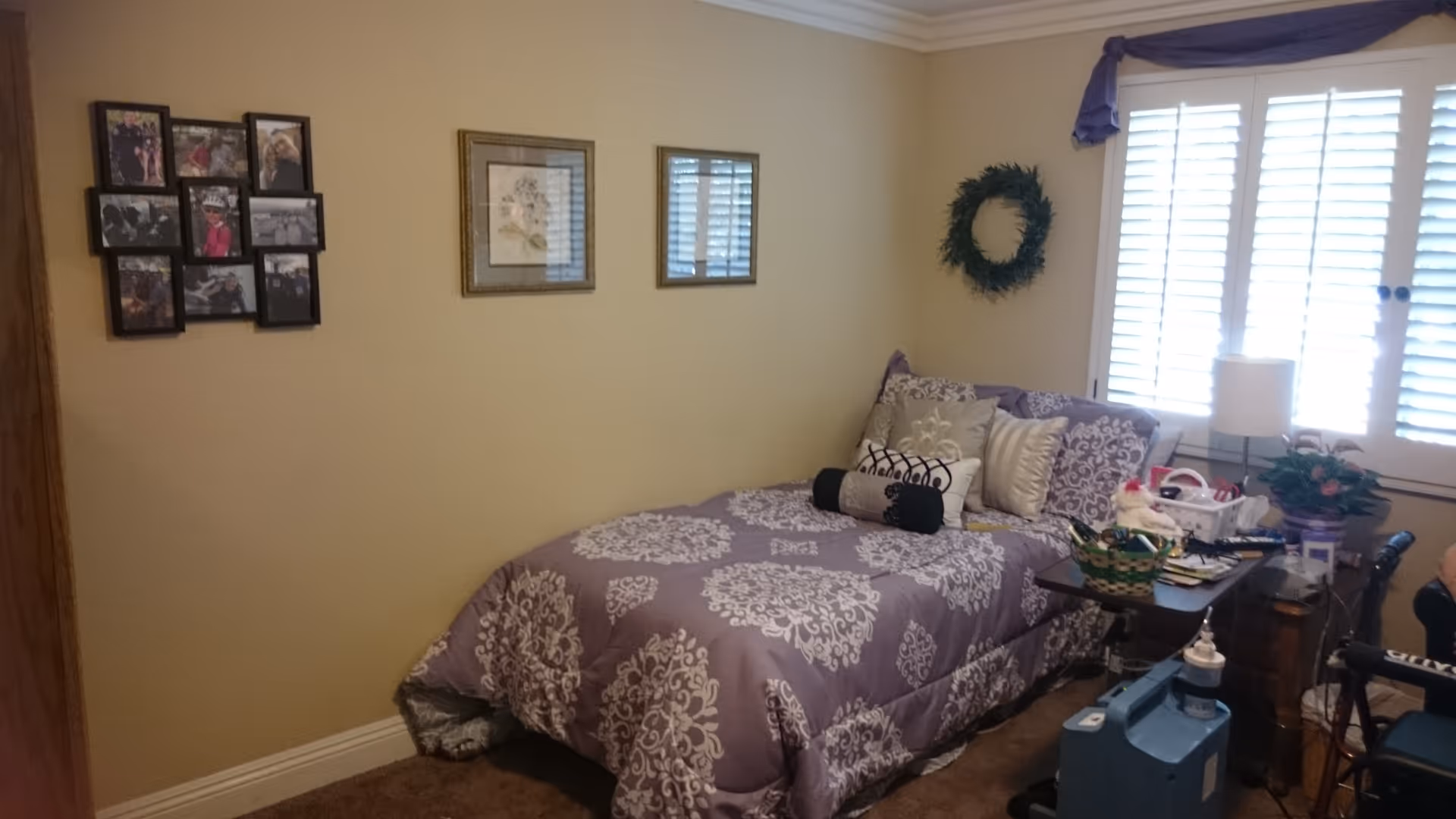 A bedroom with a single bed covered in a purple and white patterned comforter and multiple decorative pillows. The room has beige walls with three framed pictures hanging on one wall and a green wreath on another. There is a window with white plantation shutters and a purple valance. Next to the bed is a small table with various items on it, including a lamp, a basket, and medical supplies. A wheelchair is partially visible on the right side of the image.