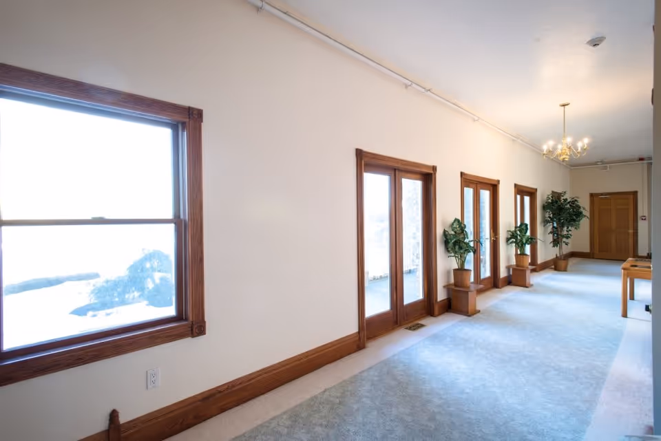 Bright carpeted interior corridor with wooden-framed windows and glass doors, potted plants, and a chandelier.