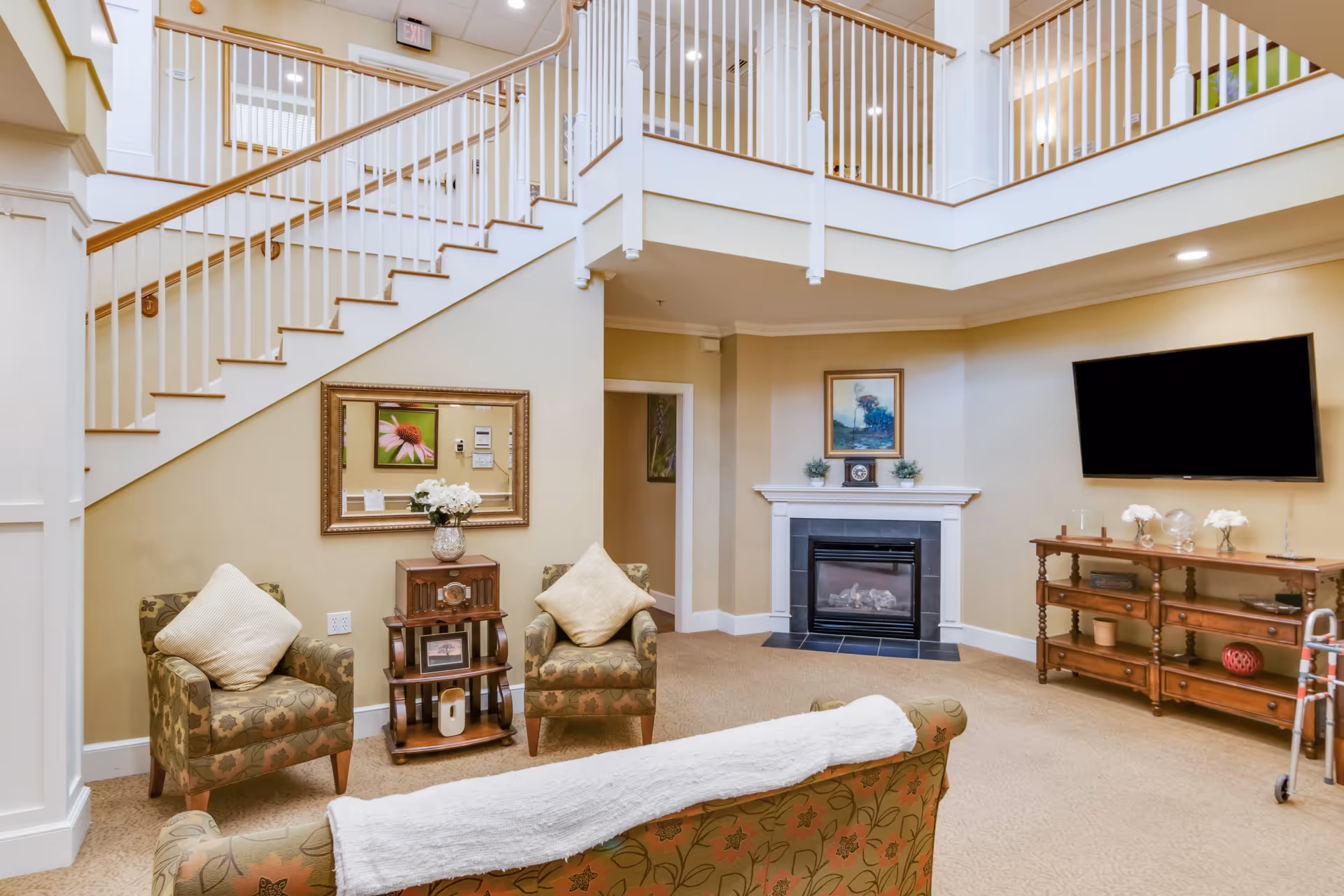 A cozy living room area in a senior living facility featuring a staircase with white railings, two patterned armchairs with cream-colored pillows, a small wooden side table with a vase of flowers, a fireplace with a framed picture above it, a flat-screen TV mounted on the wall, and a wooden console table with decorative items. The room has beige walls and carpeted flooring.