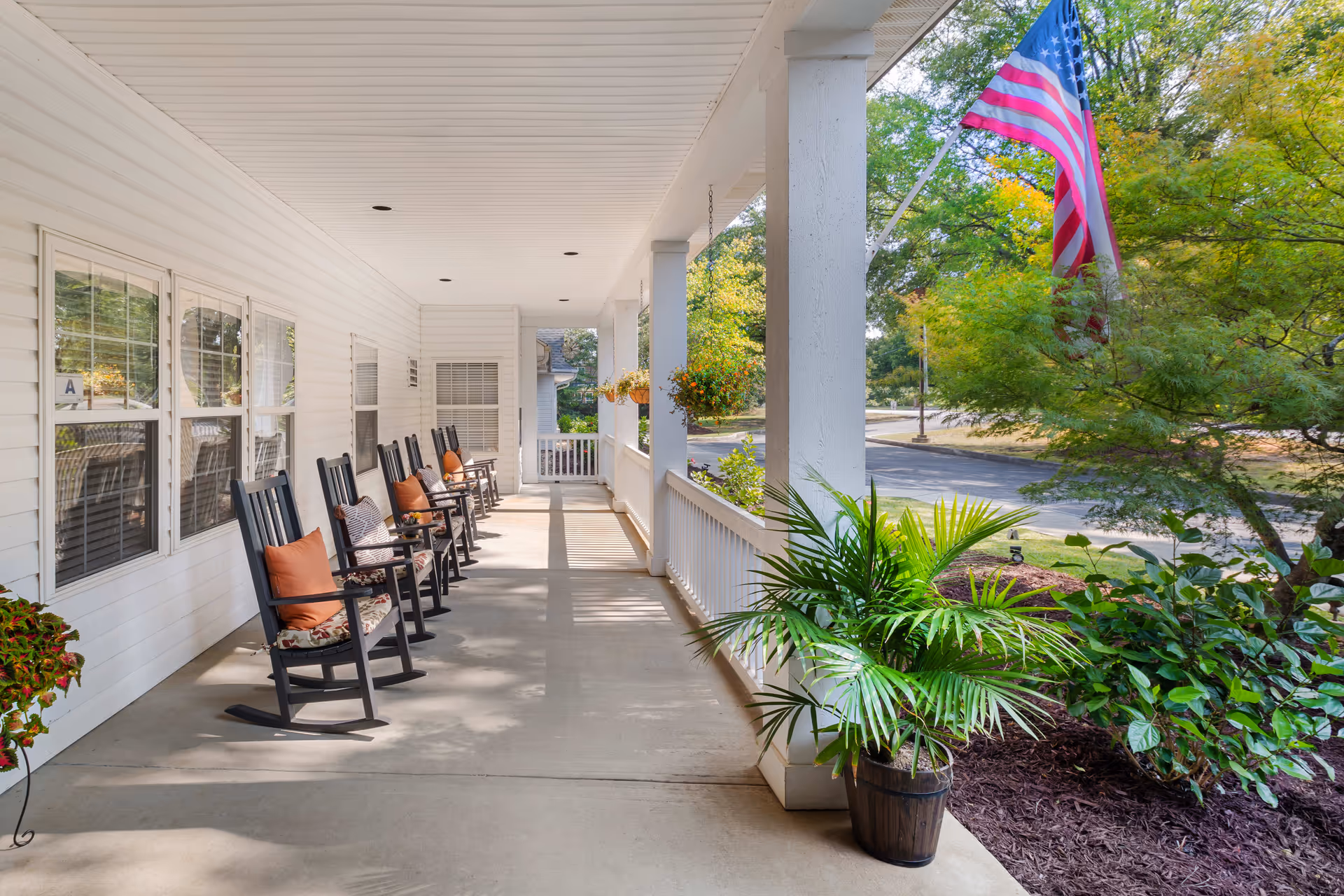 A long covered porch with several black rocking chairs lined up against the white exterior wall, each with cushions. There are hanging flower pots attached to the porch ceiling and a potted plant on the ground near the white railing. An American flag is displayed on a pole near the porch, with green trees and a road visible in the background.