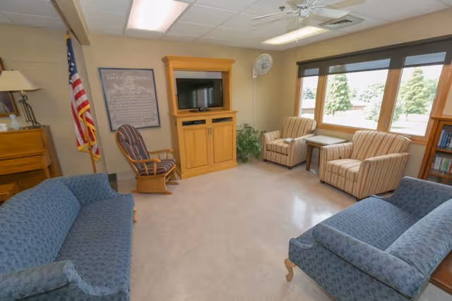 A cozy living room with two blue patterned sofas facing each other, two striped armchairs near large windows, a wooden TV cabinet with a flat-screen TV, an American flag, a wooden rocking chair, a lamp on a wooden side table, and a ceiling fan overhead.