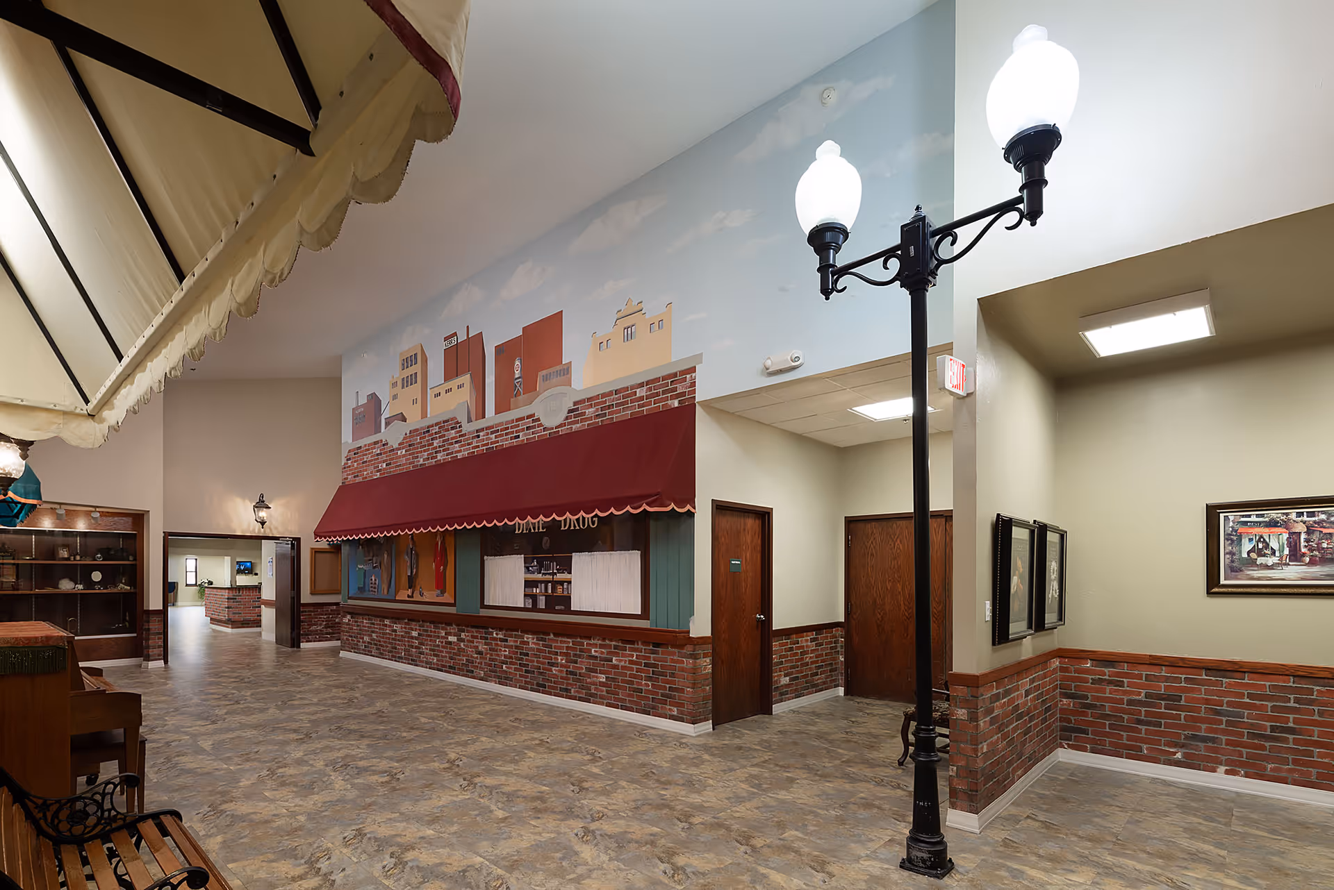 Interior view of a senior living facility hallway designed to resemble a small town street with a brick facade, awnings, and a street lamp. The walls feature a mural of buildings against a blue sky with clouds. There are wooden doors and framed pictures on the walls, with a bench and display shelves visible.