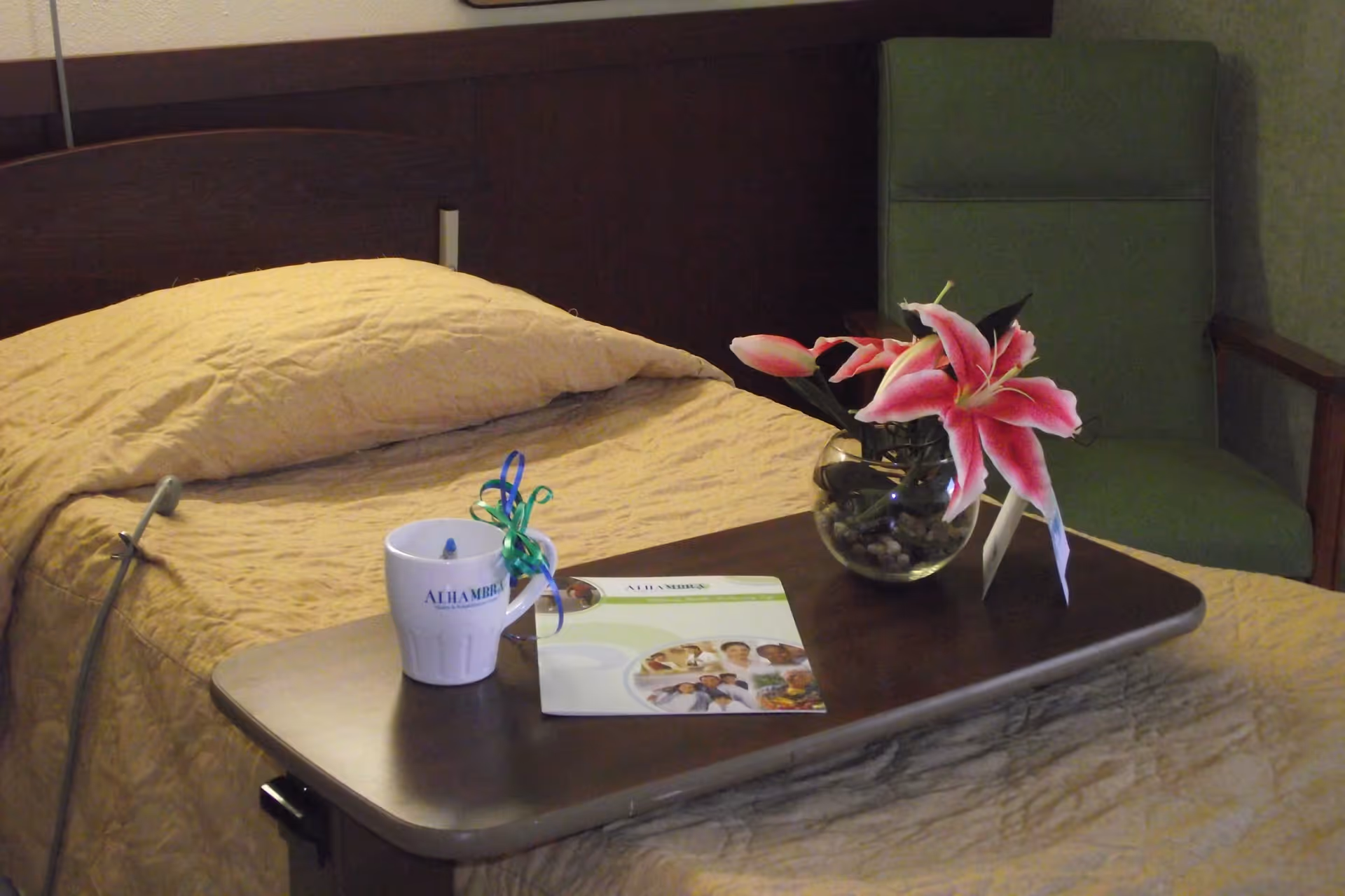 A hospital-style bed with a beige blanket and pillow, a dark wooden overbed table holding a white mug with the text 'ALHAMBRA', a brochure, and a glass vase with pink lilies. In the background, there is a green upholstered chair.