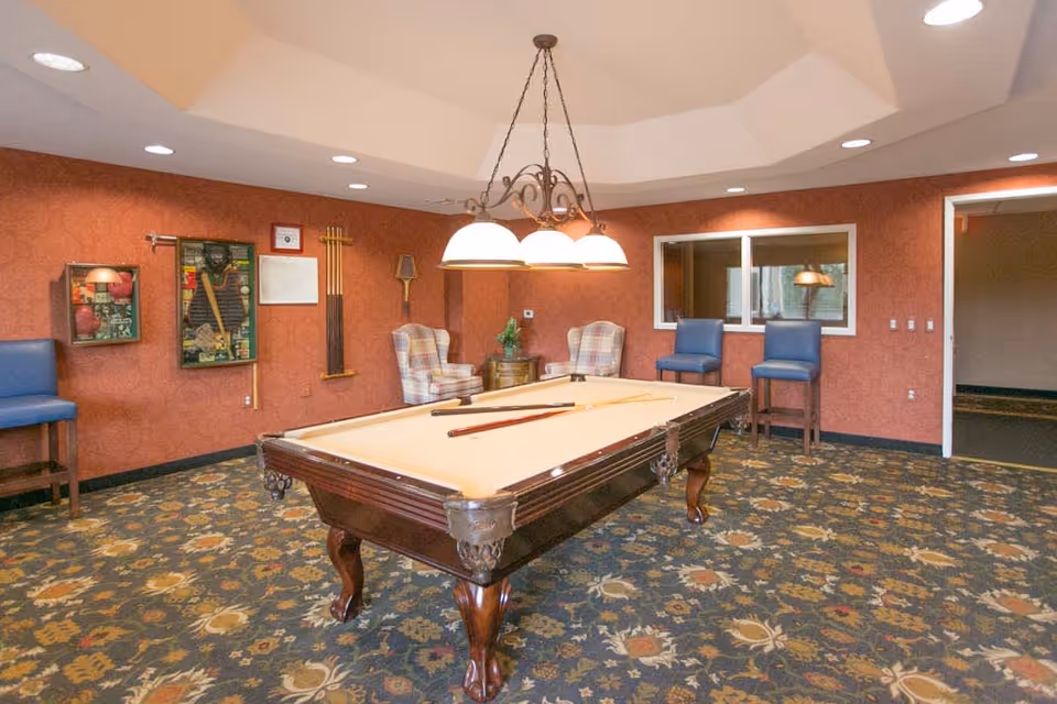 A recreational room with a pool table in the center, two pool cues resting on the table, a hanging light fixture above, patterned carpet, red textured walls, two plaid armchairs, two blue bar stools, and a window looking into another room.