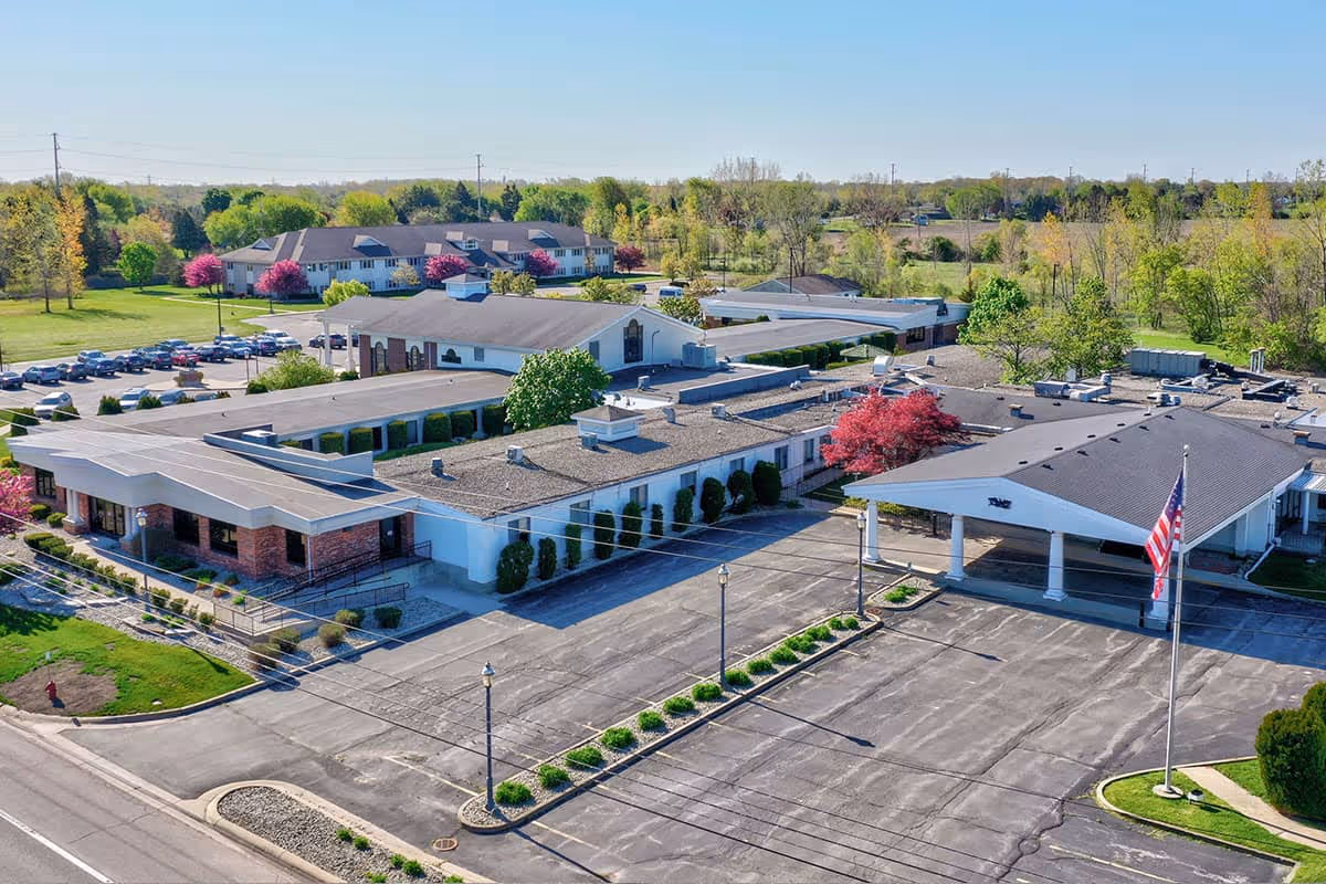 Aerial view of a single-story nursing and rehab campus with a large parking lot, entrance canopy, and surrounding trees and lawns.