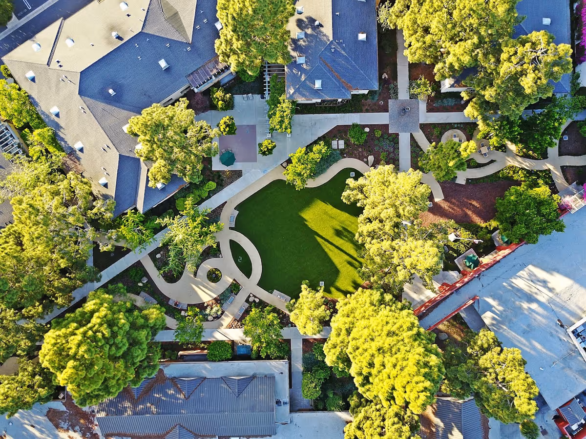 Aerial view of Vista del Monte Retirement Community showing buildings surrounded by trees, landscaped pathways, and a central green lawn area with benches and shaded seating.